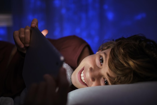 Positive preteen boy with curly hair using tablet and looking at camera in dark bedroom at home