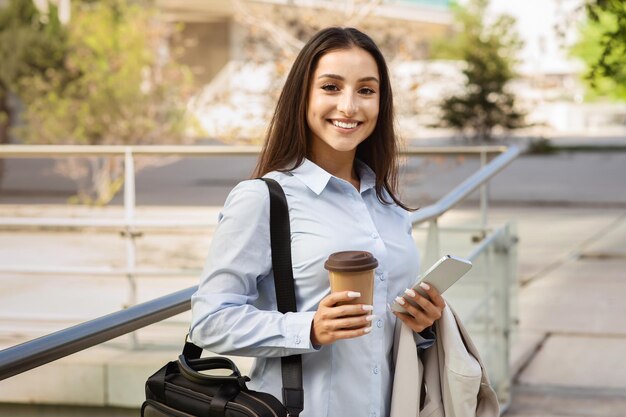 Positive pretty millennial caucasian lady manager with cup of coffee takeaway smartphone