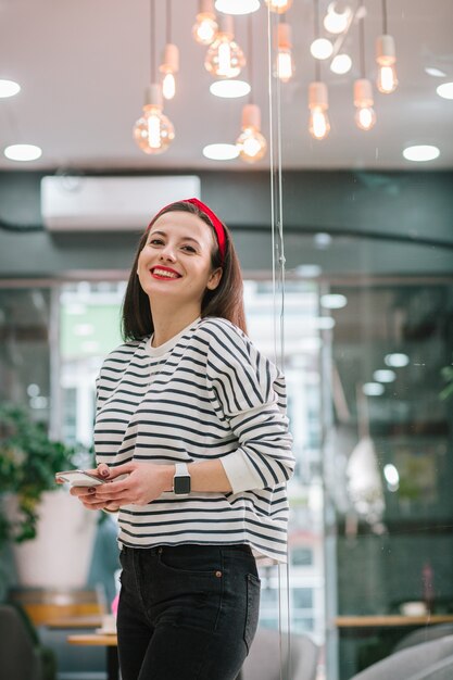 Positive pretty woman in black jeans and striped pullover standing next to the glass door with smartphone