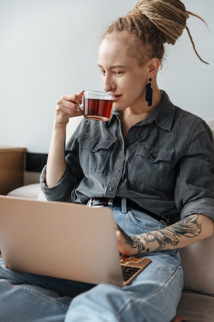 ドレッドヘアを持ち、ラップトップコンピューターを使用して屋内でお茶を飲みながらピアスをしているポジティブなかなり若い女の子。