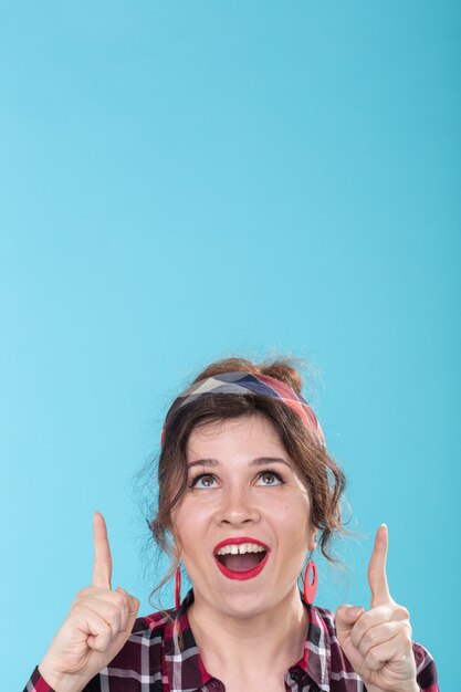 Positive pretty young woman with red earrings joyfully shows her finger upwards posing on a blue