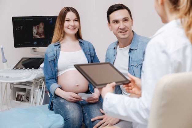 Positive qualified involved medic enjoying working hours in the hospital and using gadget while young couple enjoying appointment