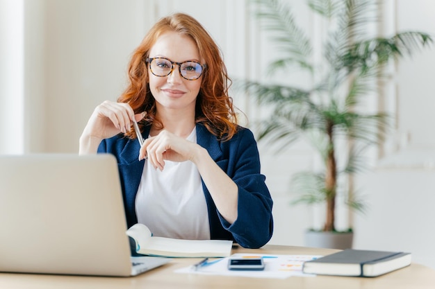 Positive red haired young female entrepreneur watches business webinar writes email makes notes in notepad wears optical glasses dressed in formal clothes works online involved in banking sphere