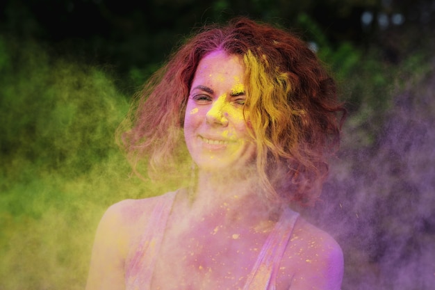 Positive redhaired model wearing white t shirt playing with purple and yellow dry Holi paint exploding around her