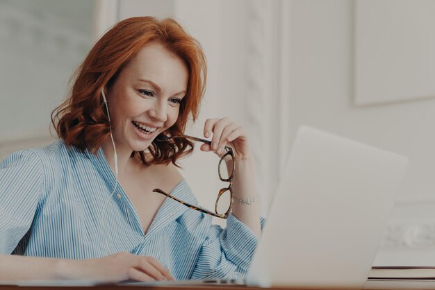 Positive redhead businesswoman participates in online conference has training class with tutor focused in display of laptop computer holds spectacles enjoys watching educational webinar