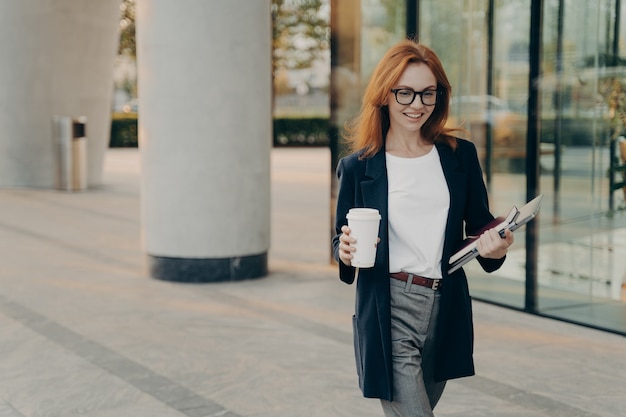 Positive redhead young European woman in elegant clothes holds notebook pen and laptop