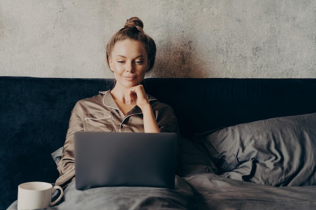 Positive relaxed young woman in pajama lying in bed and using laptop