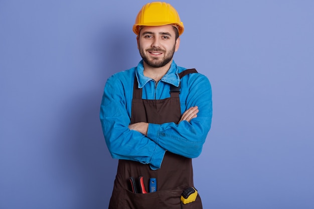 Positive repairman wears yellow protective construction helmet, shirt and brown apron, ready for house renovation.