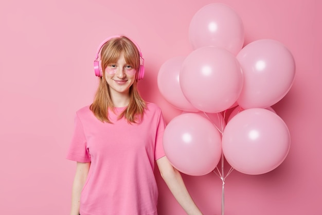 Positive rerhead fifteen years old girl listens music via\
headphones dressed in casual t shirt holds bunch of inflated\
balloons prepare for holiday applies beauty patches under eyes\
poses indoor