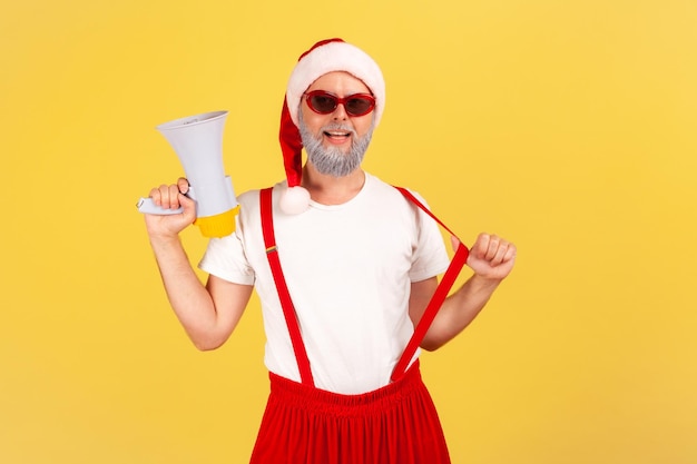 Positive self confident adult man in santa claus costume holding megaphone and looking at camera ready to organize your holiday party Indoor studio shot isolated on yellow background