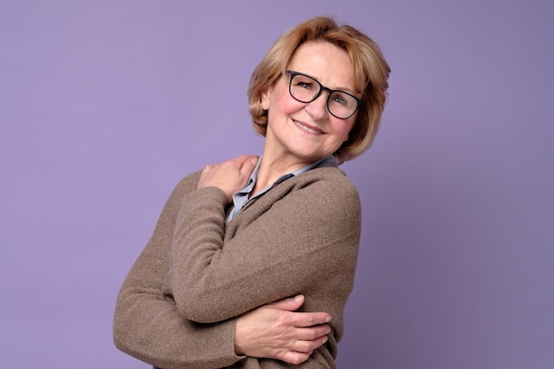 Positive senior caucasian woman in glasses smiling keeping arms around herself