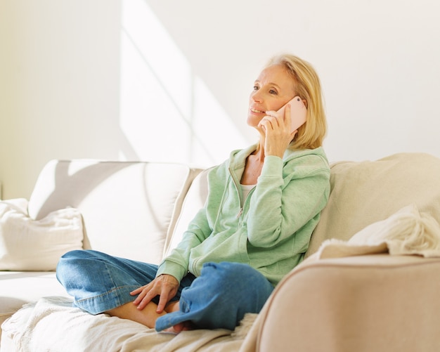 Positive senior woman enjoying pleasant call on mobile phone while relaxing on sofa at home