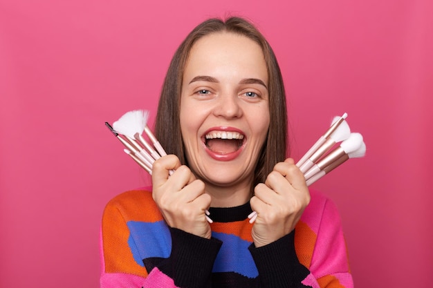 Positive sincere woman makeup artist with natural beauty wearing sweater holding cosmetic brushes to apply decorative makeup standing isolated over pink background