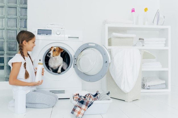 Positive small girl has fun with jack russel terrier poses on floor near washing machine in laundry room holds liquid powder busy with housework has glad expression Cozy interior in bathroom