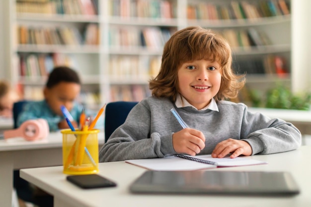 Positive smart schoolboy sitting at desk in classroom smiling at camera while writing in notebook