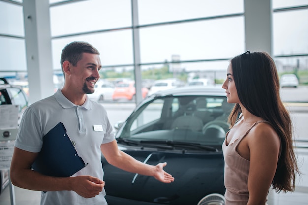 Positive and smileful man stand and point on black car. He looks at young woman. She is listening to him. Model is calm and peaceful.