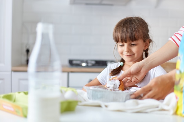 Bambina sorridente positiva con due trecce in piedi vicino al tavolo, in attesa di sua madre che le dà una cottura calda e gustosa, ragazza del bambino che indossa una maglietta bianca in posa in cucina.