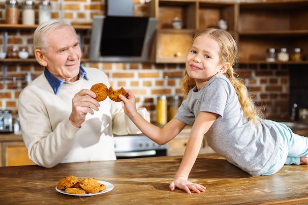 Positive smiling little girl eating cookies with her grandfather while resting in the kitchen