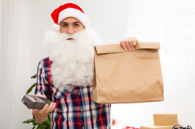 Positive smiling male courier in uniform and santa claus hat holding payment terminal and paper shopping bags, easy express order and delivery. Indoor studio