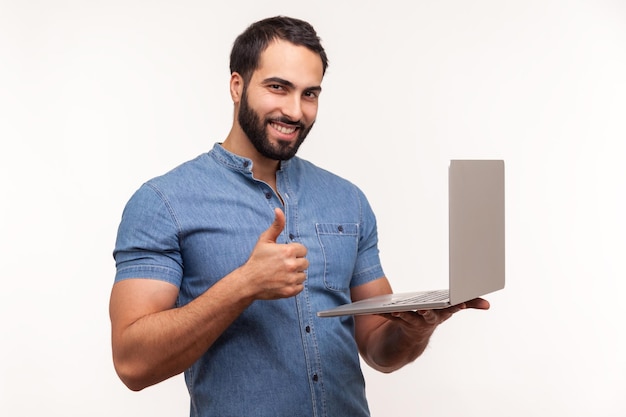 Positive smiling man with beard holding laptop in hand showing thumbs up gesture blogger likes posts in social networks recommending to followers Indoor studio shot isolated on white background