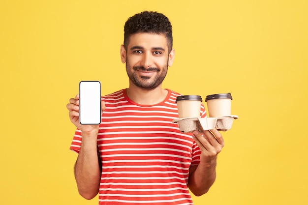 Positive smiling man with beard in striped t-shirt holding in hands empty display smartphone and holder with coffee paper cups, online order. Indoor studio shot isolated on yellow background