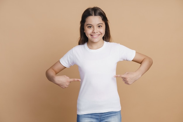 ポジティブで笑顔の10代の少女は、広告の1か月である白いTシャツを指しています。茶色の背景の少女