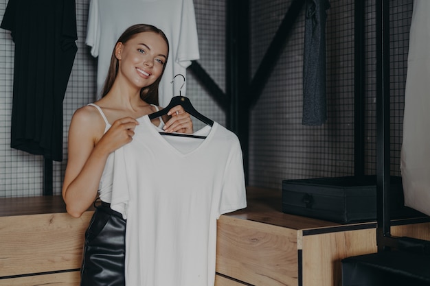 Positive smiling woman dresses for special occasion holds white dress on hangers poses over home wardrobe