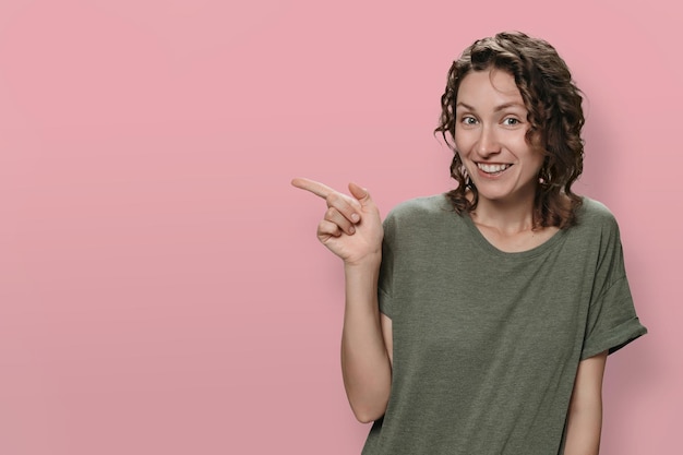 Positive smiling woman indicates with index finger at blank copy space isolated on pink background