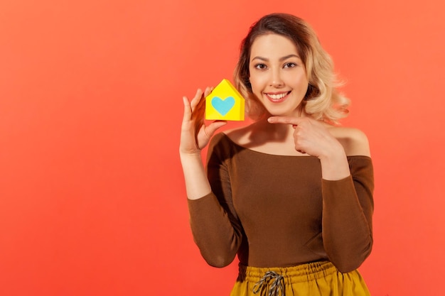 Positive smiling woman pointing finger at little yellow paper\
house with drawn heart she holding building orphanage indoor studio\
shot isolated on orange background