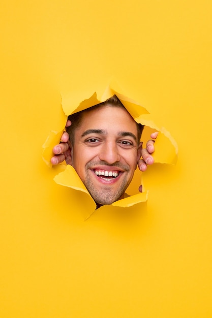 Positive smiling young bearded male looking at camera through hole in ripped yellow paper