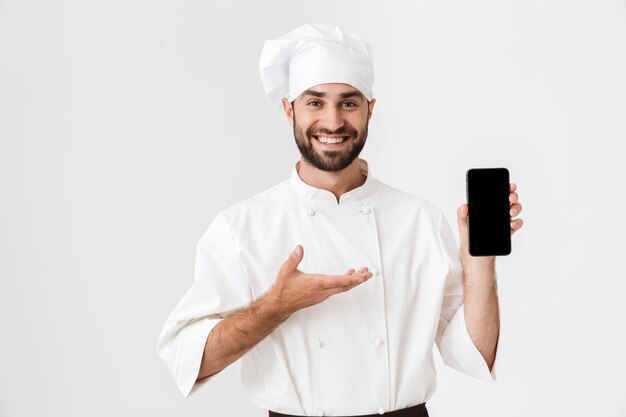 Giovane chef sorridente positivo che posa in uniforme che tiene il telefono cellulare che mostra display vuoto.