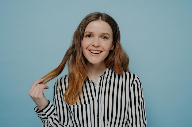 Positive smiling young girl in stripy black and white blouse demonstrating curiosity and amusement while holding strand of wavy brown hair posing over light blue background