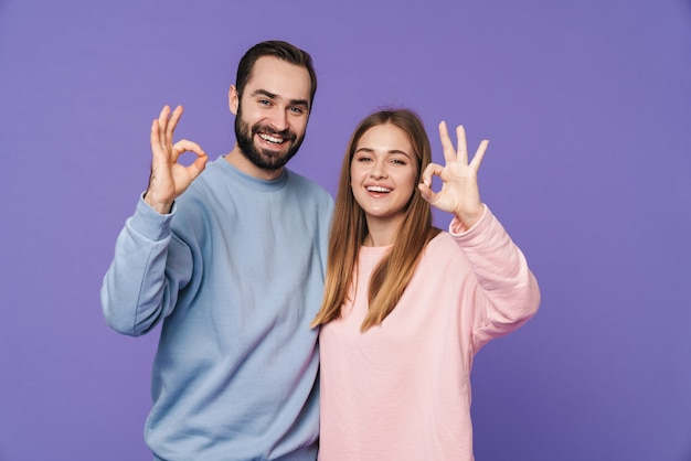 Una giovane coppia amorosa sorridente positiva isolata sopra la parete viola che mostra il gesto giusto.