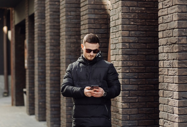 Positive smiling young male in stylish clothes standing outside alone near wall of urban building and speaking on mobile phone