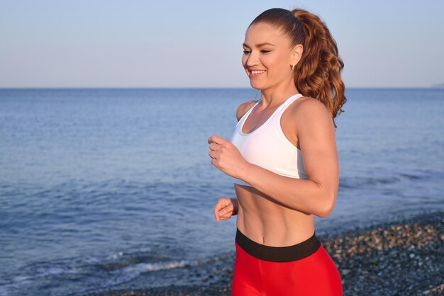 Positive sportive woman on a summer morning jogging on the beach in red leggings on the background of the sea coast close uo