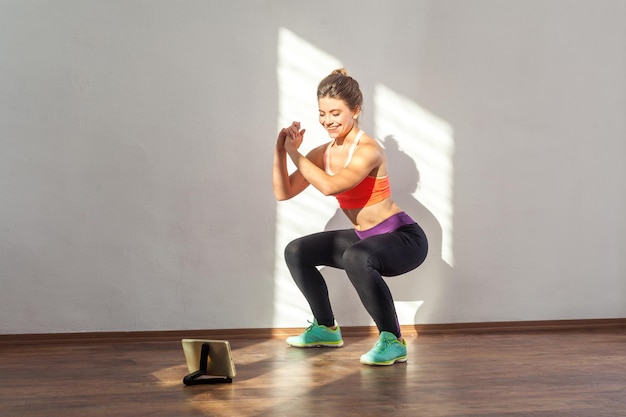 Foto donna sportiva positiva con acconciatura da panino e abbigliamento sportivo attillato che fa esercizio di posizione accovacciata mentre si guarda il video di allenamento su tablet in studio al coperto illuminato dalla luce del sole dalla finestra