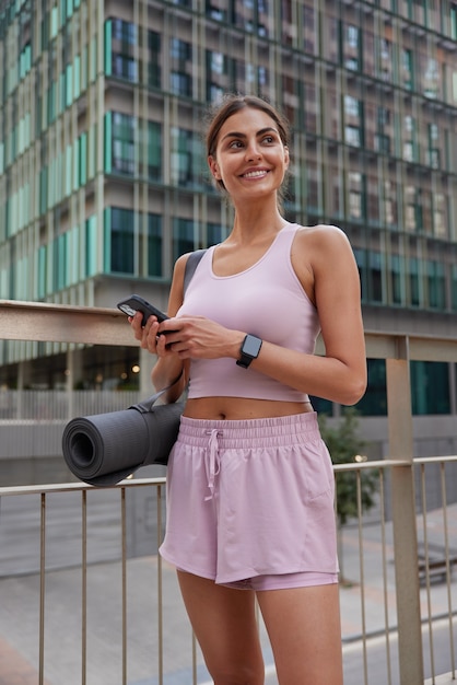 Positive sportswoman in cropped pink to and shorts holds mobile phone chats online carries karemat resturns from aerobics session has smile on face poses on modern cityscrapers outdoors