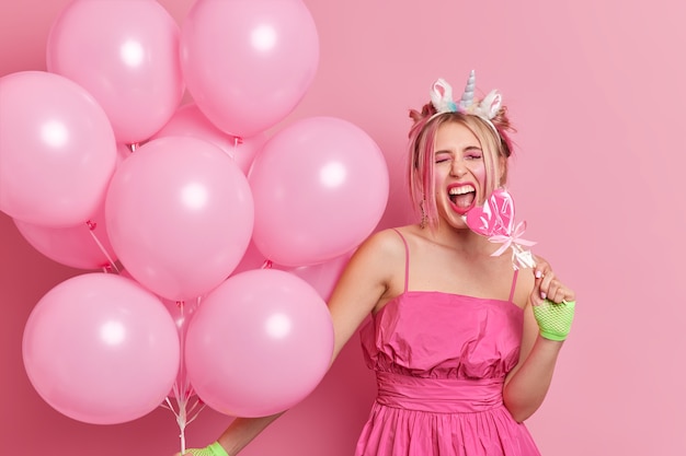 Positive stylish birthday girl has fun on party holds lollipop and bunch of air balloons wears festive dress unicorn headband 