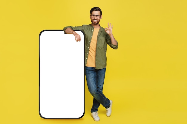 Positive stylish man showing okay gesture standing next to huge smartphone with blank screen advertisement mockup