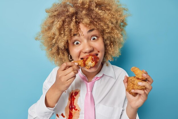 Positive surprised curly haired woman eats delicious nuggets\
with ketchup enjoys eating junk food wears white formal shirt with\
tie isolated over blue background unhealthy nutrition concept