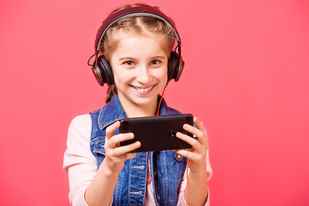 Positive teen girl listening to music in headphones and holding smartphone while looking at camera p