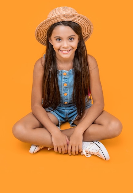 Positive teen girl in straw hat sitting on yellow background