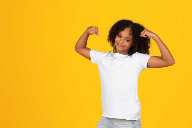 Positive teenage black pupil girl in white tshirt shows biceps muscles