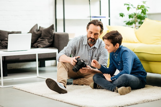 Positive teenage boy sitting on the carpet next to the sofa and looking at the camera in hands of his father