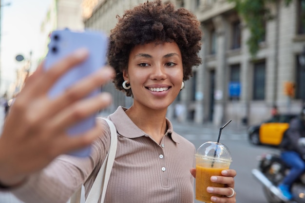 Positive teenage girl strolls on road drinks cocktail clicks selfie photos on cellular has happy expression dressed casually poses outdoors outdoor makes picture of herself