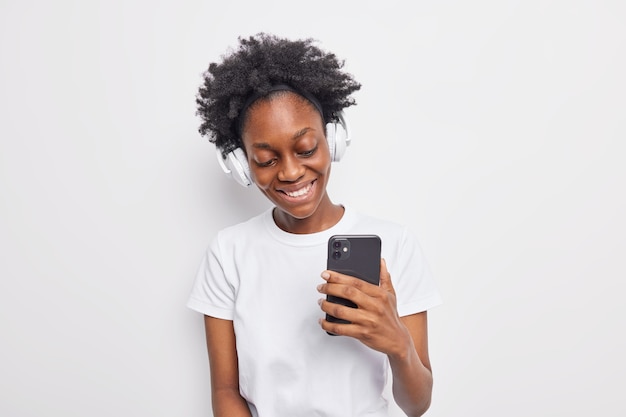 Positive teenage girl with Afro hair listens to  favorite song from playlist holds modern smartphone uses wireless headphones 