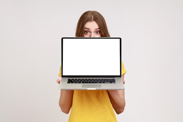 Позитивная неизвестная женщина в желтой футболке прячет половину лица за ноутбуком с белым пустым дисплеем и смотрит в камеру большими удивленными глазами. Снимок в помещении, изолированный на сером фоне