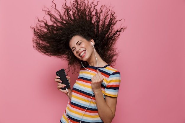 Photo positive woman 20s with shaking hair singing while holding smartphone and listening to music via headphones isolated on pink