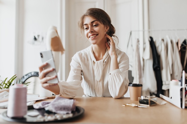 Positive woman in beige blouse holds phone and sits in office