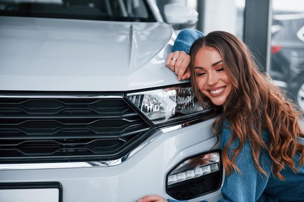 Позитивная женщина в синей рубашке обнимает свой новый автомобиль в автосалоне